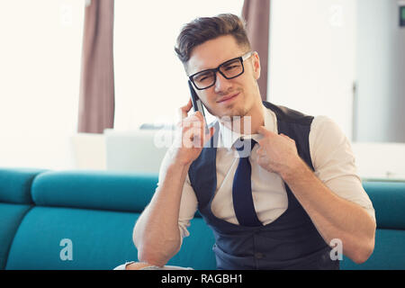 Man in glasses looking anxious while having phone call and frowning on couch in modern apartment Stock Photo