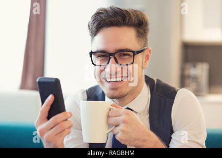 Young smiling man in glasses surfing phone and drinking coffee sitting on couch looking at camera Stock Photo