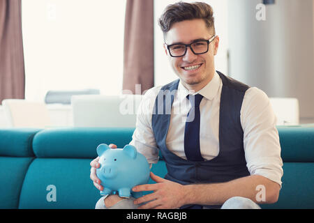 Happy formal man in glasses holding blue piggy bank sitting on sofa in new modern apartment smiling at camera Stock Photo