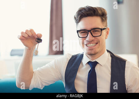 Young smiling man in eyeglasses holding key from new house sitting on couch Stock Photo