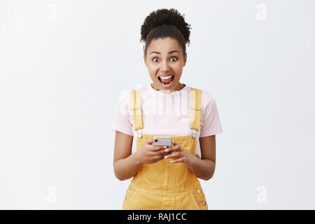Womans creaming from emotions, receiving invitation to famous party. Portrait of surprised and amazed good-looking dark-skinned girl in yellow overalls, yelling at camera and holding smartphone Stock Photo