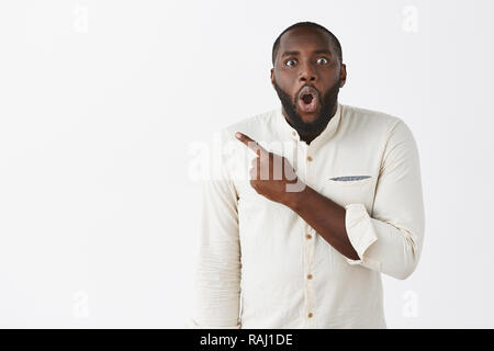 Studio shot of impressed and surprised curious adult African American in white trendy shirt, pointing at upper left corner, gasping, holding breath, being speechless from amazement over grey wall Stock Photo