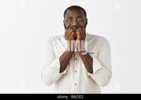 Man gets frightened hearing scary stories of friend. Scared intense and nervous attractive African American with short haircut in white shirt, biting fingernails and staring terrified at camera Stock Photo