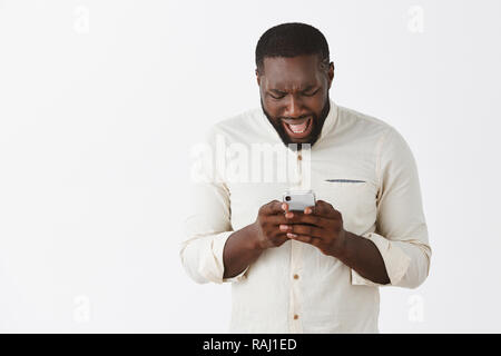 Disappointed upset dark-skinned male model in white shirt, frowning and complaining while reading disappointing message in smartphone, holding device with both hands and looking at screen Stock Photo