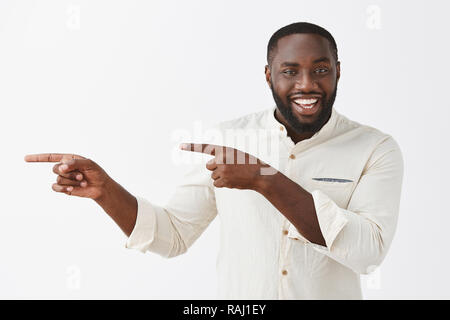 Satisfied successful and happy dark-skinned male model with beard and short haircut in white formal shirt pointing with both hands left and smiling broadly, advicing great place to eat out Stock Photo