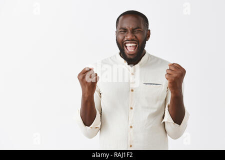 Yes, let us celebrate my success. Portrait of joyful thrilled and amused handsome African American in white shirt raising clenched fists and yelling from happiness, triumphing from victory or win Stock Photo