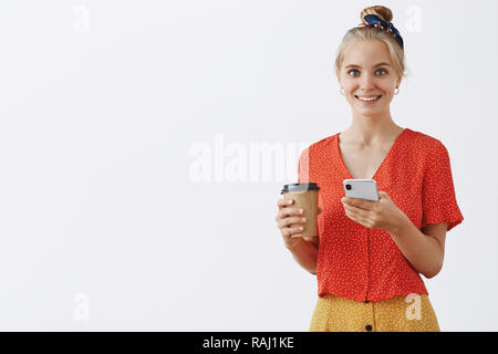 Waist-up shot of chamring blonde woman with combed hair holding paper cup of coffee and smartphone smiling joyfully at camera having break in afternoon, scrolling news in internet and drinking tea Stock Photo