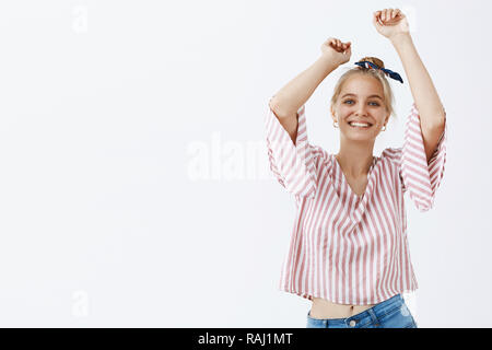 Wanna dance under summer rain. Portrait of stylish and joyful emotive fair-haired woman with combed hair in bun wearing trendy striped blouse raising hands and smiling joyfully having fun Stock Photo