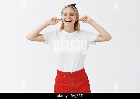 Girl feeling relieved plugging ears with index fingers hearing no sound or noise smiling broadly with satisfaction, closing eyes and standing over gray background in headband and trendy skirt Stock Photo