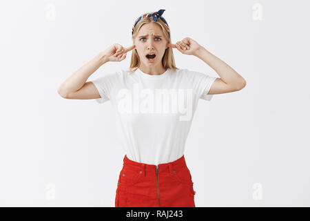 Turn off this rock and turn my songs. Displeased bothered attractive caucasian sister with blond hair in trendy outfit, frowning and gasping, plugging index fingers in ears. being annoyed with noise Stock Photo