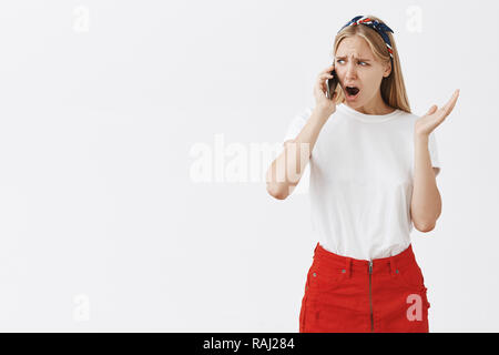 Studio shot of displeased moody girlfriend with blond hair frowning and compaining while talking on smartphone, gesturing and grimacing being displeased during argument with friend Stock Photo