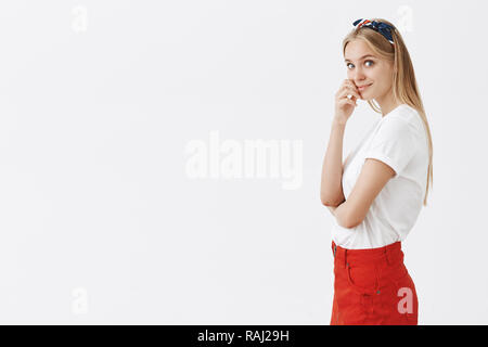 Perfect day to dream and create. Curious good-looking clever female with fair hair in headband and red skirt, standing in profile at turning at camera, touching lip, smiling flirty and cute Stock Photo