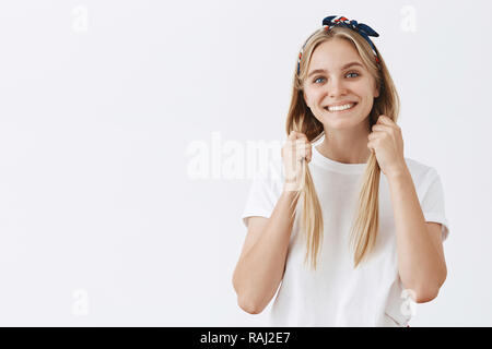 she is still little girl in heart charming cute fair haired woman in headband pulling hair down with both hands and smiling joyfully being in playful and joyful mood having fun over grey wall raj2e7