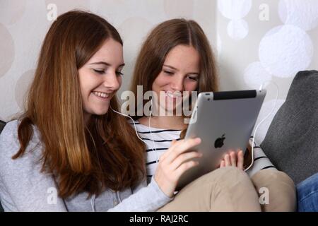 Two girls sitting on a couch, listening with earphones to music from an iPad, tablet computer Stock Photo