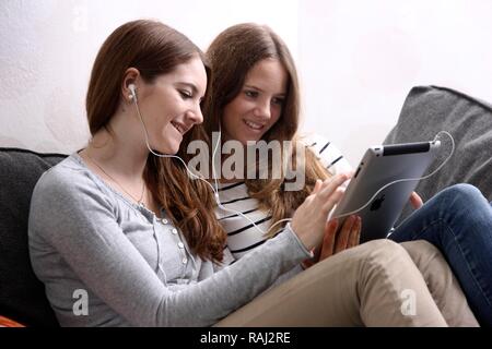 Two girls sitting on a couch, listening with earphones to music from an iPad, tablet computer Stock Photo