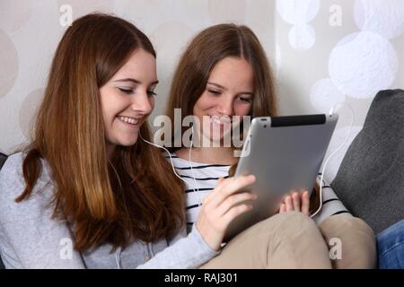 Two girls sitting on a sofa listening to music with headphones, holding an iPad, tablet computer Stock Photo
