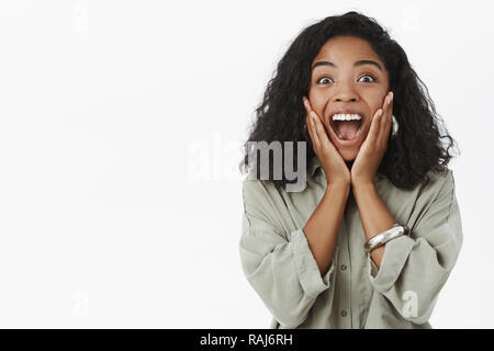 Portrait of upbeat enthusiastic and delighted surprised dark-skinned girlfriend with curly hairstyle yelling from amazement and joy receiving awesome surprise touching cheeks from happiness Stock Photo