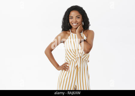 Dark-skinned woman feeling shy posing in front of boyfriend in new overalls being charming and marvellous preparing go out on date touching cheek gently being timid and feminine posing over gray wall Stock Photo