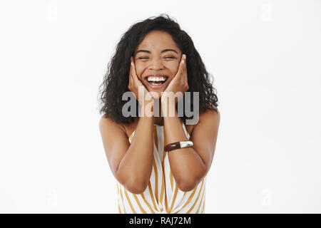 Happy joyful friendly-looking young african american woman feeling delighted and fresh after washing face holding hands on cheeks and smiling broadly having great result applying facial skincare mask Stock Photo