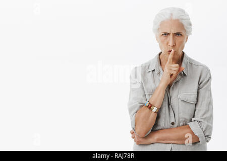 Granny dislikes when grandchild swearing. Portrait of serious-looking dissatisfied and angry energized elderly woman with white hair frowning shushing at camera with index finger over mouth Stock Photo