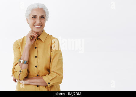 Age is nonsense. Portrait of happy charismatic stylish senior woman with white hair in trendy yellow coat posing with hand above chin and crossed arm smiling from delight and joy over gray background Stock Photo