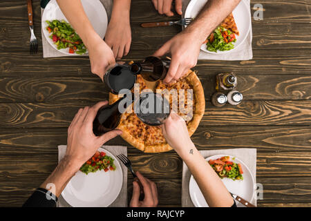 cropped view of friends toasting while having dinner with pizza and salads at wooden table Stock Photo