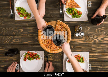 partial view of friends toasting while having dinner with pizza and salads at wooden table Stock Photo
