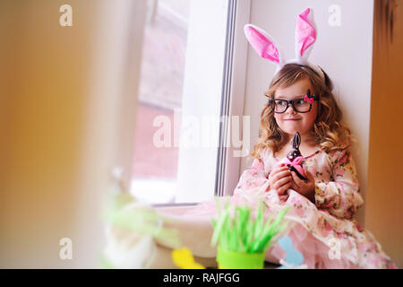 baby beautiful girl in a smart dress with rabbit ears on her head holding a chocolate Easter bunny Stock Photo