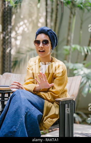 Fashion portrait of a young and beautiful Malay Asian woman (Muslim) in a turban and sunglasses. She is relaxing in a trendy cafe. Stock Photo