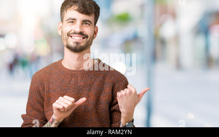 Young handsome man wearing winter sweater over isolated background Pointing to the back behind with hand and thumbs up, smiling confident Stock Photo