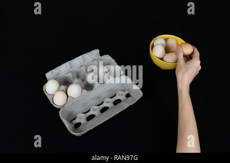 Female hand taking one egg from yellow bowl. Cardboard egg box with three eggs on black felt background. Stock Photo