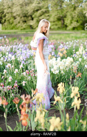 Young european girl wearing white dress standing near irises on garden. Stock Photo