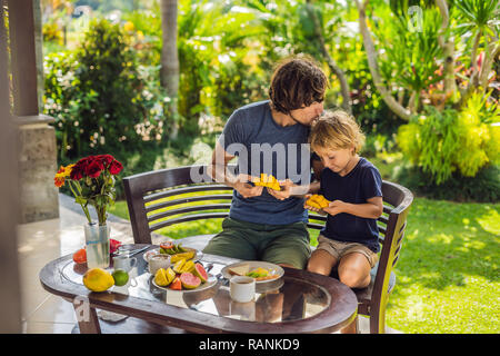 family of two eating nicely served breakfast outside handsome young man pouring some coffee and his cute son eating delicious fruit at breakfast time Stock Photo