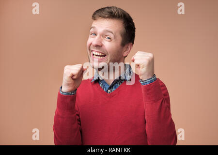 Excited surprised young man in red sweater keeping clenched fists at his faces, shocked Stock Photo