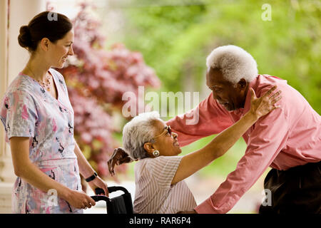 Nurse looks on as senior woman in wheelchair reaches up to embrace senior man who is reaching down to hug her. Stock Photo