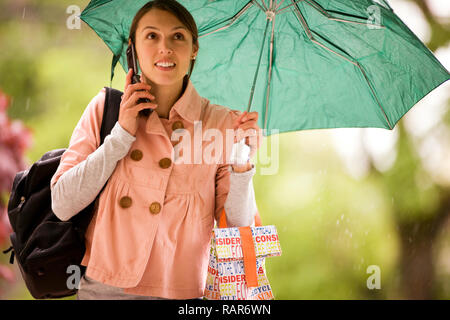Young woman uses her cellphone as she shelters under her umbrella while she walks in the rain. Stock Photo