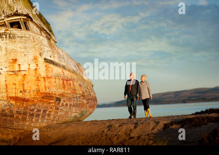 Senior couple walking on the beach hand-in-hand next to an obsolete boat. Stock Photo