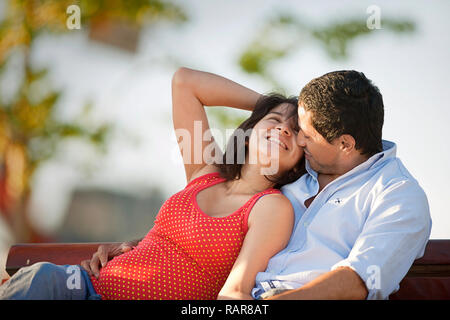 Smiling young woman lies back in the arms of a young man as they sit together on a park bench. Stock Photo