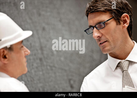 Businessman in a hardhat talks with a male colleague wearing eyeglasses. Stock Photo