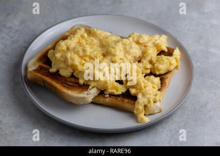Scrambled eggs on two pieces of toast on a white plate with a grey rim on a simple grey and white marbled background Stock Photo
