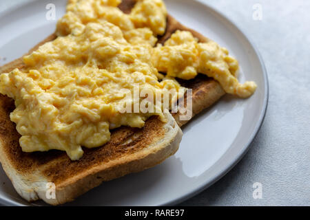 Close up of scrambled eggs on two pieces of toast on a white plate with a grey rim on a simple grey and white marbled background Stock Photo