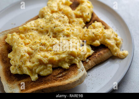 Close up of scrambled eggs with black pepper on two pieces of toast on a white plate with a grey rim on a simple grey and white marbled background Stock Photo