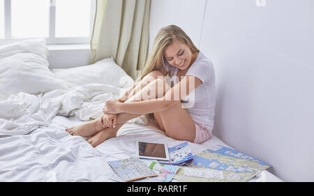 Happy young girl in bed using phone, tablet and maps to search for travel. Stock Photo