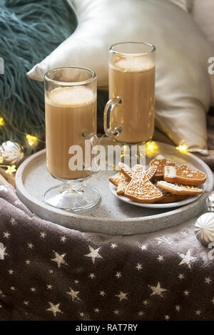 Homemade latte in glass cups, served with gingerbread on the background of plaid, pillows and garlands. Stock Photo