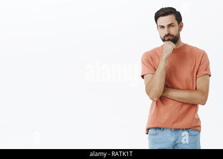 Guy having hesitations, thinking. Portrait of unsure and thoughtful attractive nice bearded guy with dark hair squinting doubtful and uncertain holding fist on chin as making choice over gray wall Stock Photo