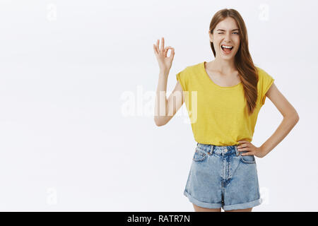 I got everything under control. Friendly charming caucasian girl in yellow t-shirt and shorts holding hand on hip winking joyfully and smiling showing okay sign, feeling excellent over gray background Stock Photo