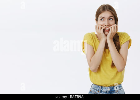 Waist-up shot of concerned nervous and anxious cute young timid woman in yellow t-shirt biting fingernails looking scared at upper left corner frightened, overreacting posing against white background Stock Photo