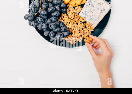 View from above woman's hand taking nut from plate with cheese, grapes, crackers and walnuts on white wooden table. Stock Photo