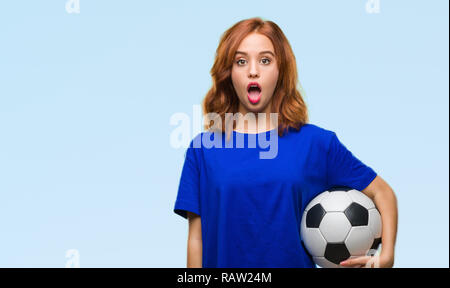 Young beautiful woman over isolated background holding soccer football ball scared in shock with a surprise face, afraid and excited with fear express Stock Photo