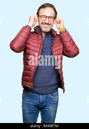 Middle age handsome man wearing glasses and winter coat Smiling pulling ears with fingers, funny gesture. Audition problem Stock Photo